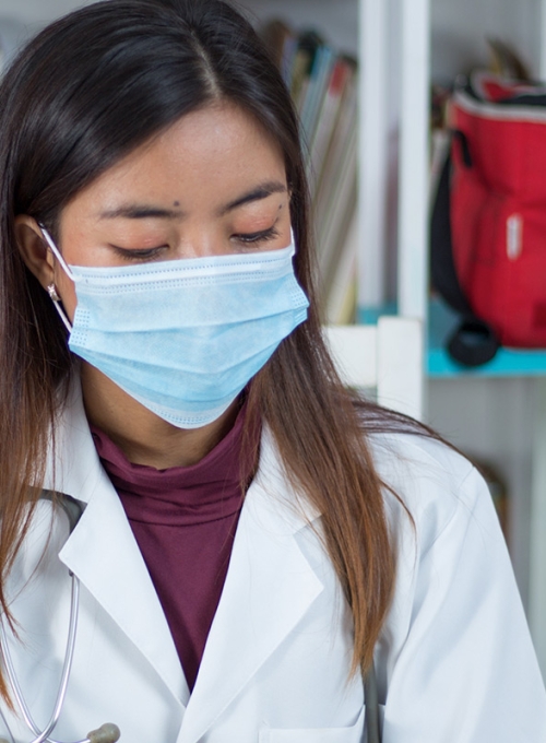Southeast Asian female doctor from Malaysia wearing her uniform and a sanitary mask in a library By Sam Salaubie/Wirestock